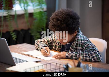 Studente africano contemporaneo in casualwear facendo note in copybook mentre seduto al tavolo davanti al computer portatile e facendo casa assegnazione Foto Stock