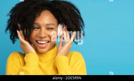 Sorridente donna da corsa mista in giallo jumper toccare le cuffie wireless, ascoltare la musica, ha buon umore, si inanchia e guarda lo spazio vuoto per l'advertis Foto Stock
