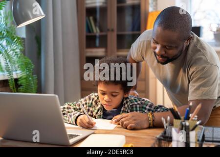 Studente africano in casualwear prendere appunti in copybook, mentre suo padre si piega sul tavolo, indicando la pagina e spiegando qualcosa a lui Foto Stock