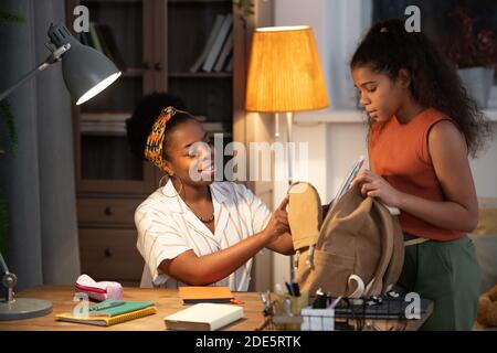 Felice giovane donna africana in casualwear aiutando la sua figlia adolescente con zaino da imballaggio per la scuola al mattino mentre si è seduti per tabella Foto Stock
