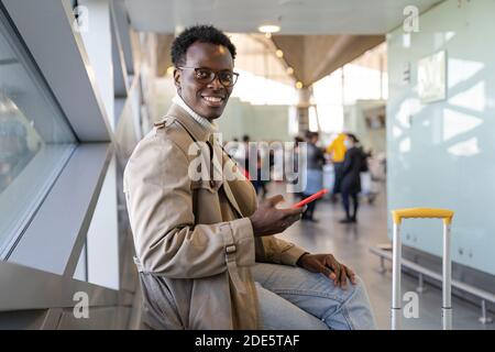 Positivo viaggiatore afro-americano uomo in trench cappotto seduto in aeroporto terminal o stazione ferroviaria, utilizzando il telefono cellulare, in attesa di volo e imbarco Foto Stock