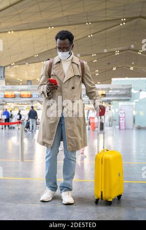 Ritratto dell'uomo viaggiatore afro-americano con valigia utilizzando il telefono, indossare la maschera protettiva per il viso durante l'epidemia di virus, coronavirus, covid-19 pandemia w Foto Stock