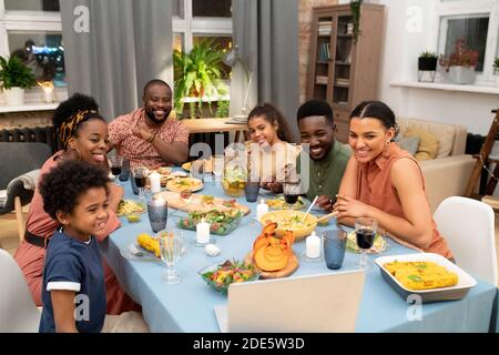 Felice famiglia africana composta da padre, madre, due figli e figlie riuniti da tavolo festivo che parla con i loro amici in video chat Foto Stock