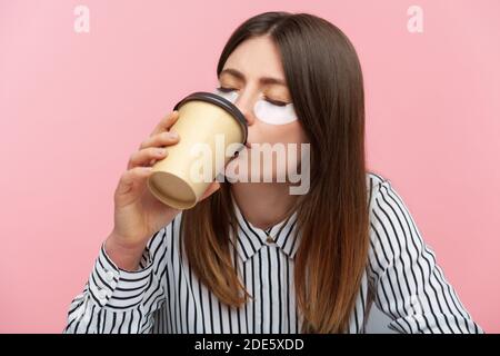 Donna sonnolenta stanca con capelli marroni in camicia a righe facendo procedura di bellezza indossare macchie per gli occhi e bere caffè da tazza di carta, mancanza di energia. IND Foto Stock