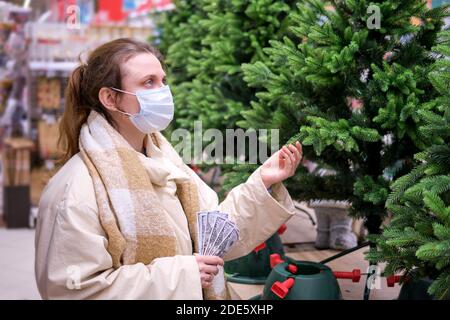 Donna in maschera di faccia con noi dollari compra l'albero di natale in negozio di articoli da regalo. Stile di vita, vita durante l'epidemia di coronavirus Foto Stock