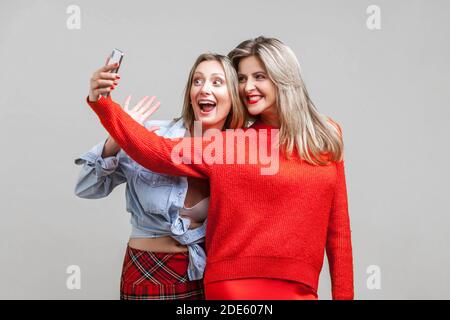 Migliori amici divertendosi. Ritratto di felici donne in abiti eleganti in piedi con sorriso toothy e tenendo selfie sul telefono insieme per godersi la vita. ind Foto Stock