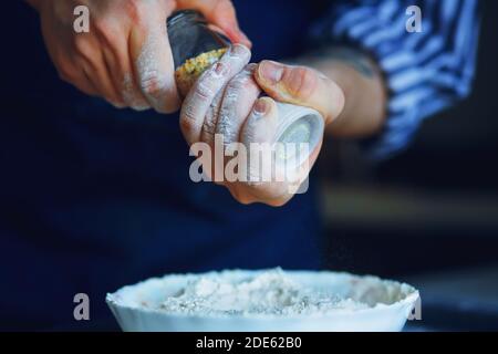 Uno chef in grembiule blu e camicia a righe tiene un bicchiere di sale shaker con condimento e lo versa in una ciotola di farina per la futura cottura. Cucina casalinga. Foto Stock