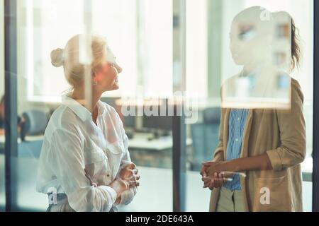 Due diverse colleghe che hanno una conversazione informale mentre si levano in piedi dentro l'ufficio moderno Foto Stock
