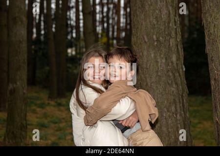 Una ragazza adolescente in un vestito leggero abbraccia il suo fratello minore. I bambini sono felici e ridono tra gli alberi della foresta. Vera infanzia e famiglia happine Foto Stock