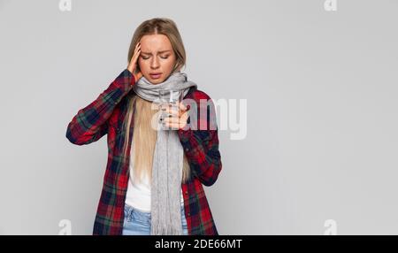 Donna in camicia controllata, sciarpa avvolta che ha mal di testa e tenendo vetro con acqua, toccando il suo tempio e gli occhi chiusi, isolato. Stagione influenzale Foto Stock