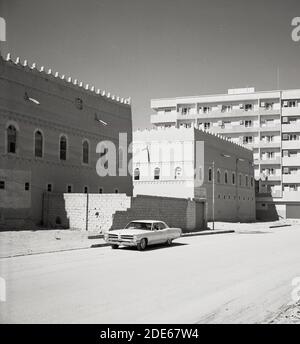 Anni '1960, storica, un'automobile americana importata parcheggiata in una strada vuota fuori da un tradizionale edificio in pietra saudita a Jeddah, in Arabia Saudita, con un moderno condominio sullo sfondo. Foto Stock