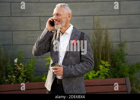 In linea. Ritratto di felice, riuscito uomo d'affari di mezza età in abito elegante sorridente mentre parla al telefono, in piedi all'aperto con una tazza di Foto Stock