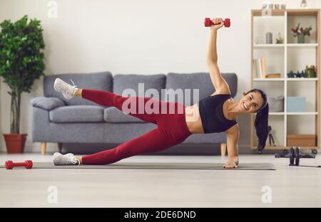 Felice giovane donna in activewear facendo fianco con manubri alzati durante l'allenamento a casa Foto Stock