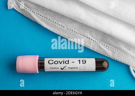 Campione di analisi del sangue positivo Covid-19 con maschera facciale protettiva isolata su sfondo blu. Concetto e background del coronavirus. Foto Stock