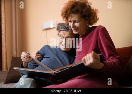 Donna anziana e sua figlia adulta guardando insieme un album fotografico sul divano in salotto, parlando gioiosamente dei ricordi. Weekend con i genitori, Foto Stock