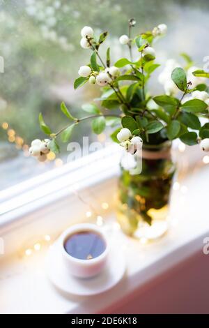 Un bacca bianca in un bouquet si erge con una tazza di caffè sul davanzale vicino alla finestra, belle luci, bokeh nella finestra Foto Stock