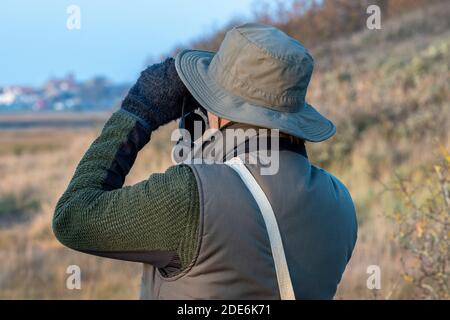 un uomo vestito di abbigliamento scuro che indossa un cappello e tiene binocoli birdwatching alla ricerca di uccelli rari. Foto Stock