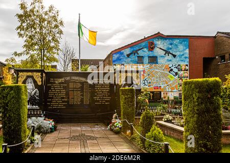 Un giardino commemorativo su Conway Street, Belfast, Irlanda del Nord, presenta una bandiera tricolore irlandese che sventola sopra un muro commemorativo nero con i nomi dei defunti; dietro, è visibile un murale che mostra una sezione di mappa stradale, medaglioni ritrattistici e due fucili incrociati. Foto Stock