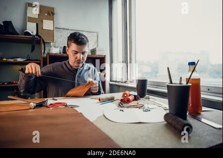 Un giovane calzolaio esperto o su misura cucina un pezzo unico di pelle in uno spazioso e luminoso studio. Foto Stock