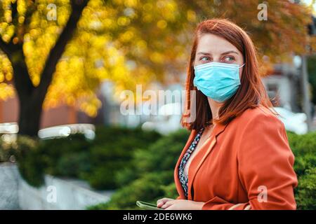 Coronavirus, covid 19 concetto. Giovane ragazza in maschera medica e tuta tiene uno smartphone nelle mani e guarda a lato sulla strada Foto Stock