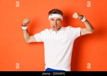 Immagine di un simpatico sportivo di mezza età con archetto e t-shirt bianchi, bicipite flessibili da mostrare, in piedi su sfondo arancione Foto Stock