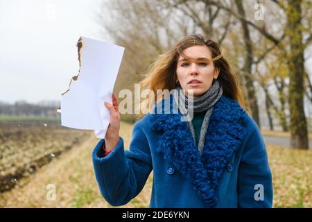 Frustrata giovane donna con make-up mascara sbavato dopo aver pianto, in cappotto blu, rimanere sul campo e tenere bruciando smolder bianco carta Foto Stock