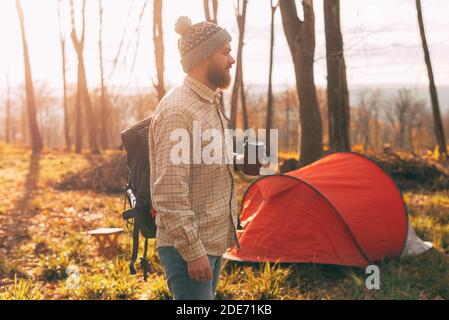 Foto di uomo bearded con tazza di caffè, indossando zaino che cammina vicino alla sua tenda in foresta. Foto Stock