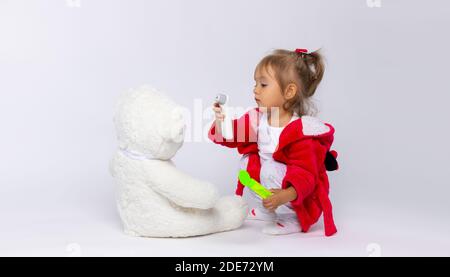 Una bambina gioca medico e misura la temperatura con un termometro senza contatto per il suo orso bianco. Foto Stock