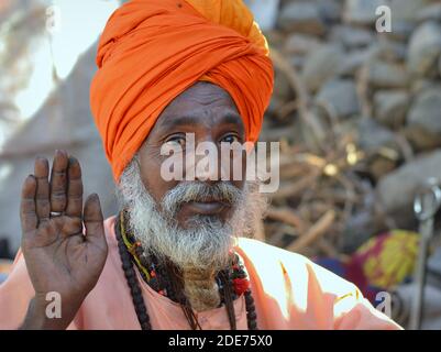 Il sadhu indiano anziano con il turbante arancione solleva la sua mano destra con la palma in avanti per dare una benedizione durante la fiera di Bhavnath (Shivratri Mela). Foto Stock