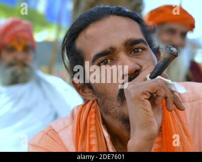 Il giovane devoto indiano religioso fuma in un tubo di chillum durante Shivratri Mela e due sadhus anziani con torbani arancioni lo guardano. Foto Stock