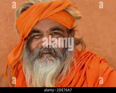 L'anziano uomo santo indù indiano con uno degli occhi (sadhu, baba) con barba piena, rivestito in arancione, si pone per la macchina fotografica davanti ad una parete marrone. Foto Stock