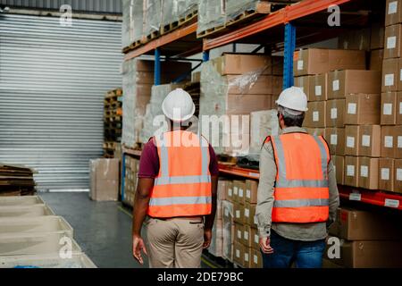 Due gara mista che lavorano insieme camminando attraverso isole di imballato pacchi in fabbrica Foto Stock