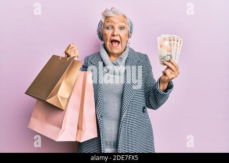 Donna senior con capelli grigi che tiene borse per la spesa e banconote inglesi in sterline che celebrano folle e stupito per il successo con gli occhi aperti che gridano eccitato. Foto Stock