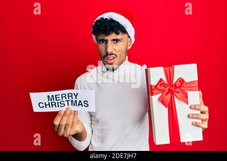 Giovane arabo bell'uomo che indossa cappello di natale tenendo allegro messaggio di natale in faccia shock, guardando scettico e sarcastico, sorpreso con aperto mout Foto Stock