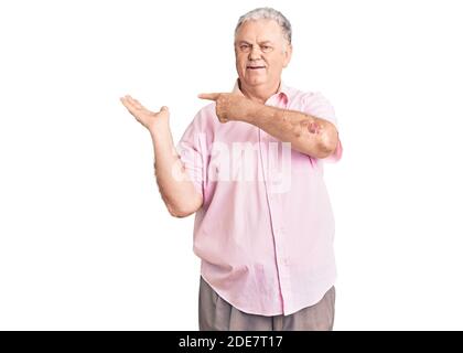 Uomo anziano dai capelli grigi che indossa abiti casual stupito e sorridente alla macchina fotografica mentre si presenta con la mano e punta con le dita. Foto Stock