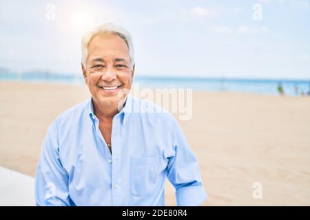 Hancosme uomo ispanico con capelli grigi sorridente felice in spiaggia, godendo vacanze in estate Foto Stock