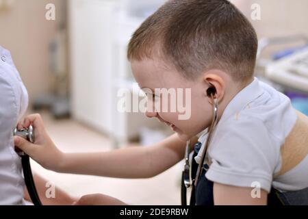 Il bambino ride in imbarazzo con le cuffie stetoscopio nelle orecchie, e applica la membrana audio testa al cappotto bianco. Comunicazione divertente Foto Stock