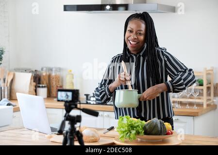 Felice donna afroamericana vlogger che trasmette video in diretta online mentre cucina in cucina a casa Foto Stock