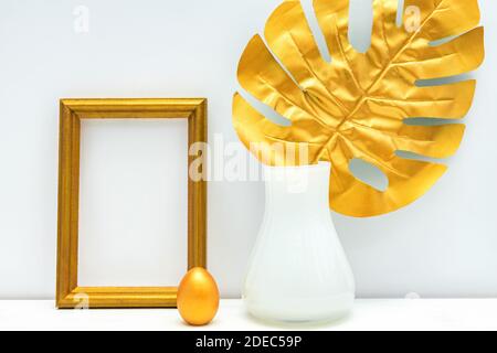 design mockup interno in oro e bianco. photoframe vuoto, uovo verniciato e foglia di monstera in vaso bianco su sfondo bianco della parete. Design elegante e lussuoso. Foto Stock