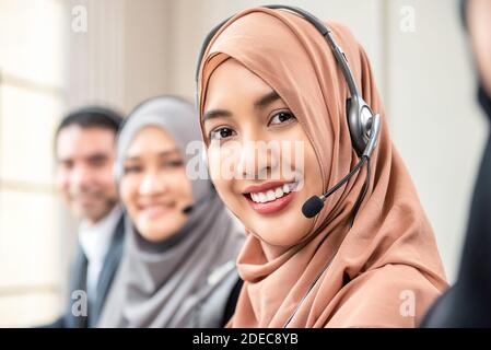 Amichevole bella donna musulmana asiatica che indossa microfono cuffia che funziona come operatore di assistenza clienti con team nell'ufficio del call center Foto Stock