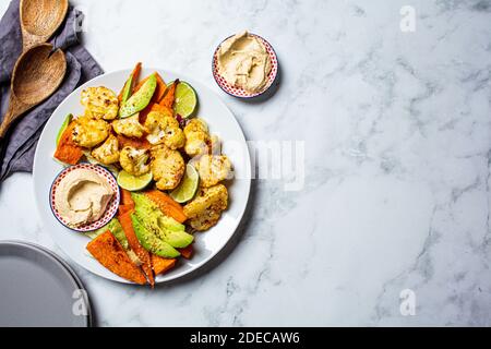 Concetto di cibo vegano. Verdure cotte con hummus in un piatto bianco, fondo di marmo bianco. Foto Stock