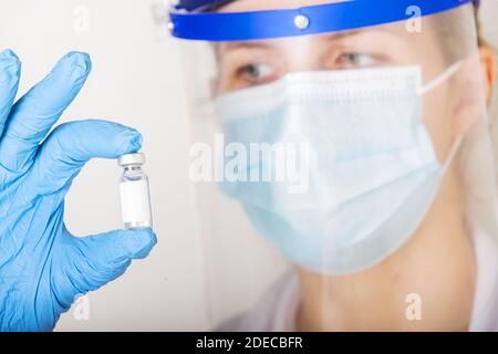 Medico con guanti blu e visiera sta tenendo una fiala di farmaco con copy-space su uno sfondo bianco. Concetto medico. Foto Stock