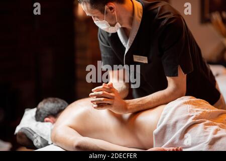 Massaggiatore professionista in maschera facciale che effettua un massaggio profondo ad un cliente maschile al salone Spa. Business durante il concetto epidemico Foto Stock