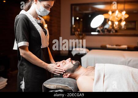 Massaggiatore femminile professionista in maschera medica che fa massaggio facciale ad un cliente maschio al salone di Spa. Business durante il concetto epidemico Foto Stock
