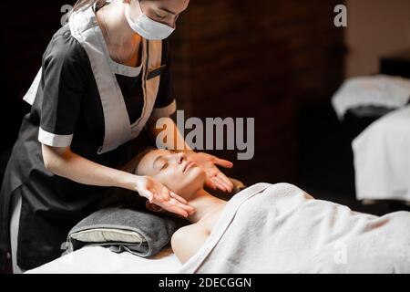 Massaggiatrice femminile professionista in maschera medica che fa massaggio facciale ad un cliente al salone di Spa. Business durante il concetto epidemico Foto Stock