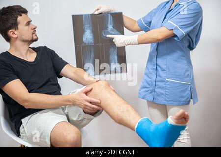 Medico femminile in un abito medico blu spiega ad un paziente di sesso maschile con una gamba rotta il risultato di una radiografia. Foto Stock