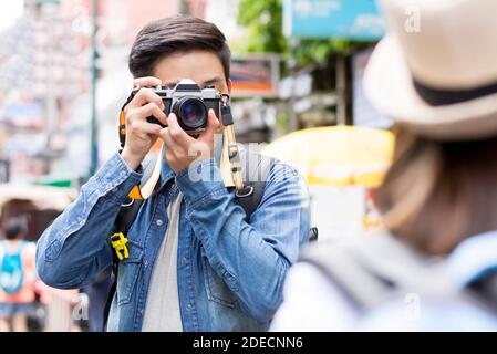 Fotografo turistico asiatico maschile in giacca blu denim scattando foto di compagno di viaggio femminile in posa in fuori città mercato di strada mentre in vacanza vis Foto Stock
