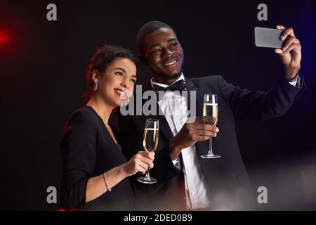 Ritratto in vita di una coppia di corse miste elegante scattando foto selfie mentre si levano in piedi contro sfondo nero durante la festa, copia spazio Foto Stock