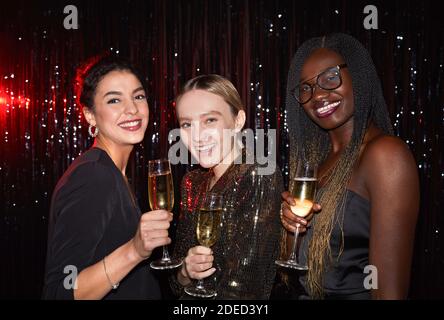 Ritratto in vita di tre eleganti giovani donne che tengono bicchieri da champagne e sorridono alla fotocamera mentre si posa su sfondo scintillante alla festa, Sho Foto Stock