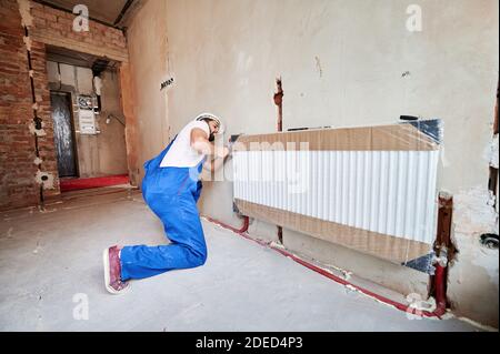 Idraulico in tute da lavoro utilizzando una chiave durante l'installazione del radiatore di riscaldamento nella stanza. Uomo in casco installazione impianto di riscaldamento in appartamento. Concetto di installazione di radiatori, lavori idraulici, ristrutturazione domestica Foto Stock